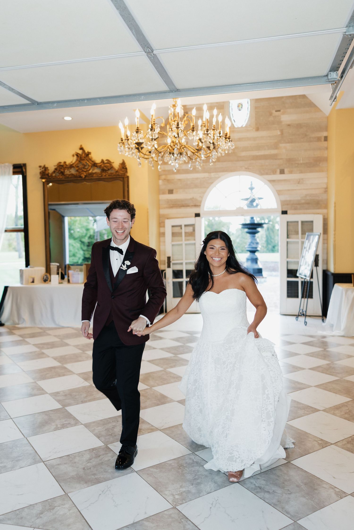 Smiling newlyweds holding hands entering a grand ballroom with chandelier and checkered floors.