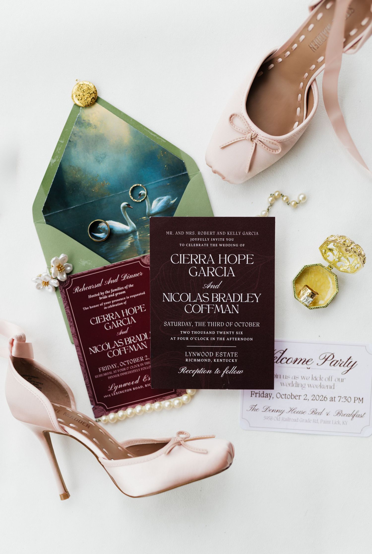 Elegant wedding flat lay with burgundy invitations, pearl shoes, pink heels, gold rings, and a swan-lined envelope on white.
