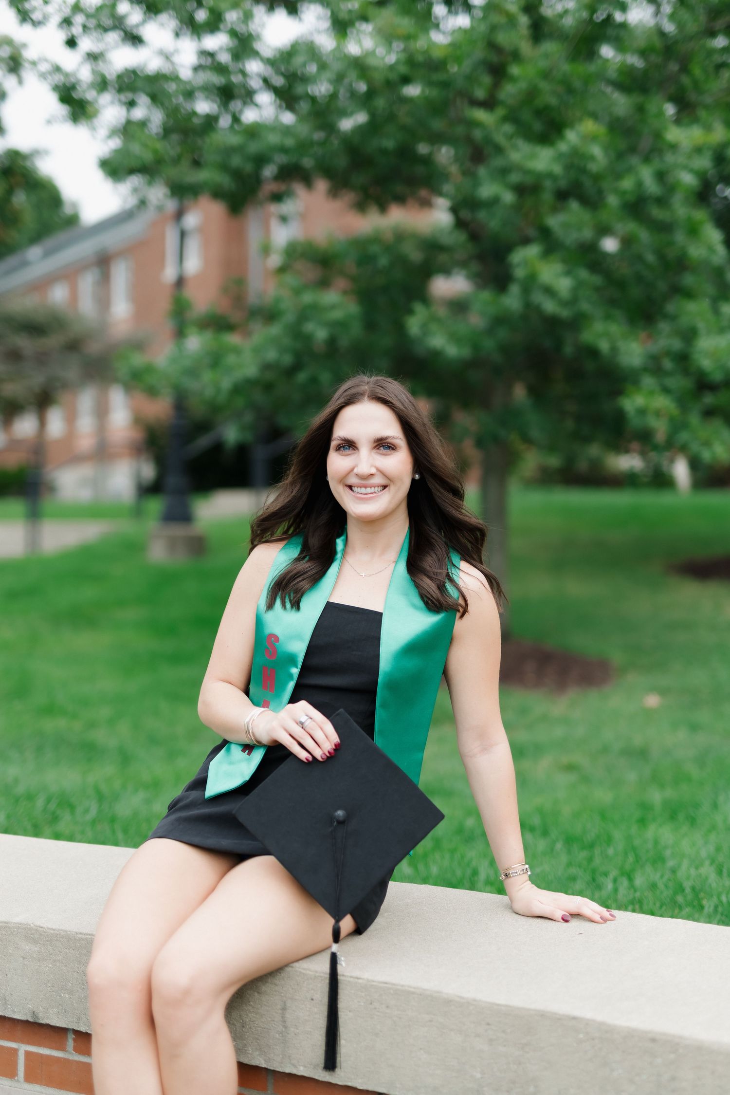 Smiling college graduate in black dress with green stole holds cap while sitting outdoors on campus.