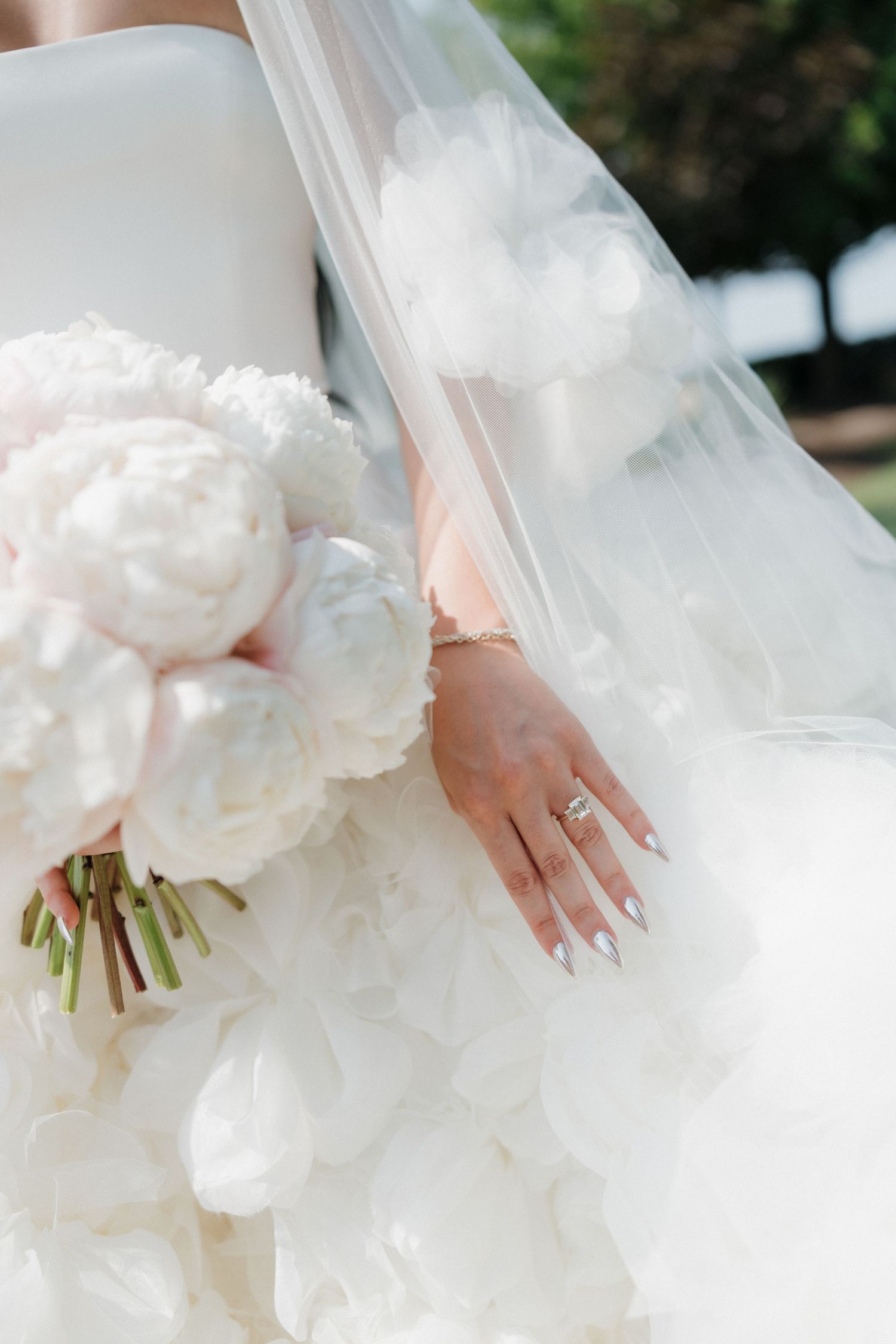 Bride holding a lush white peony bouquet, wearing a diamond ring and silver nails, with a flowing tulle veil and ruffled gown.