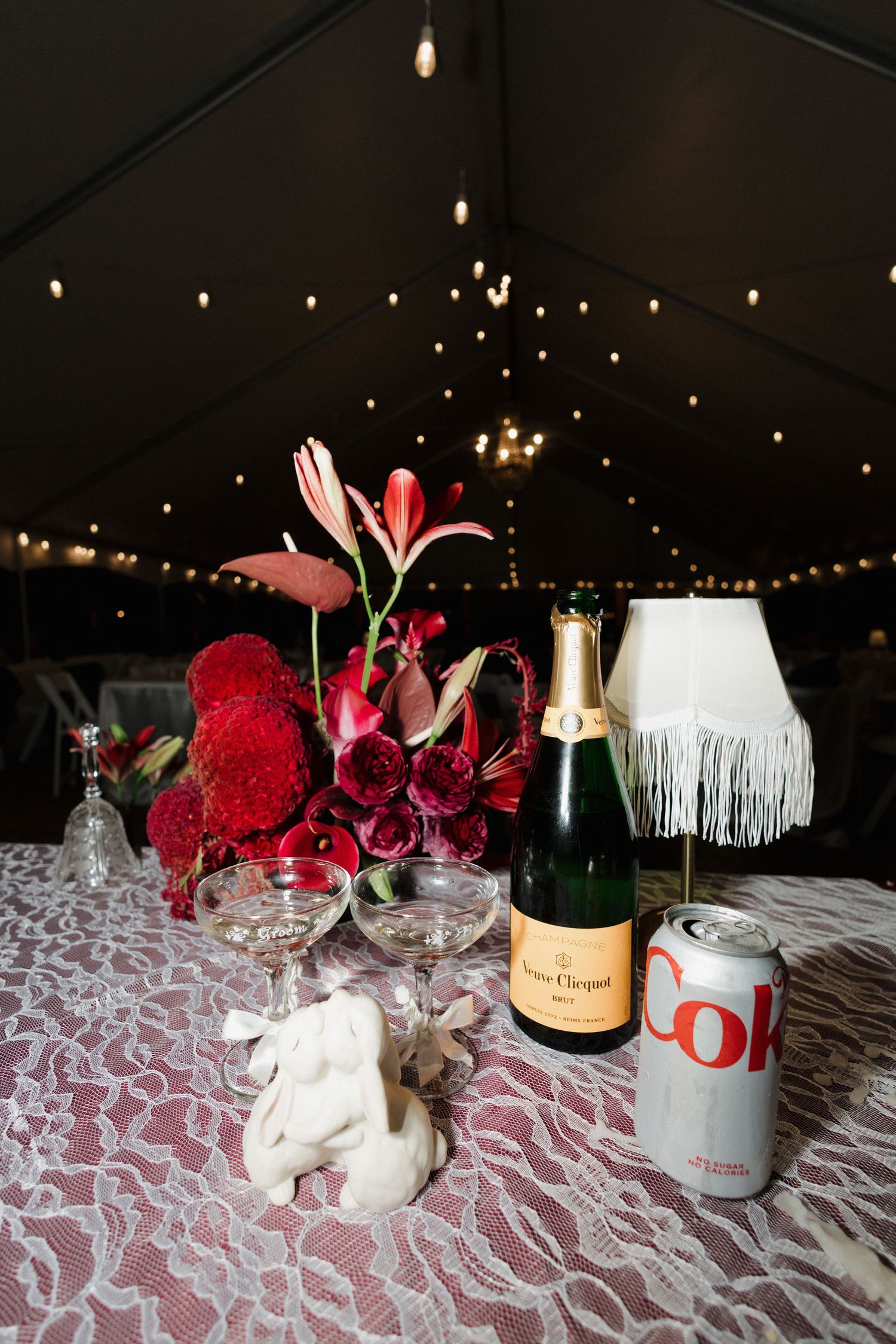 Veuve Clicquot champagne bottle with Diet Coke can, crystal glasses, red floral centerpiece, and fringe lamp on lace tablecloth.