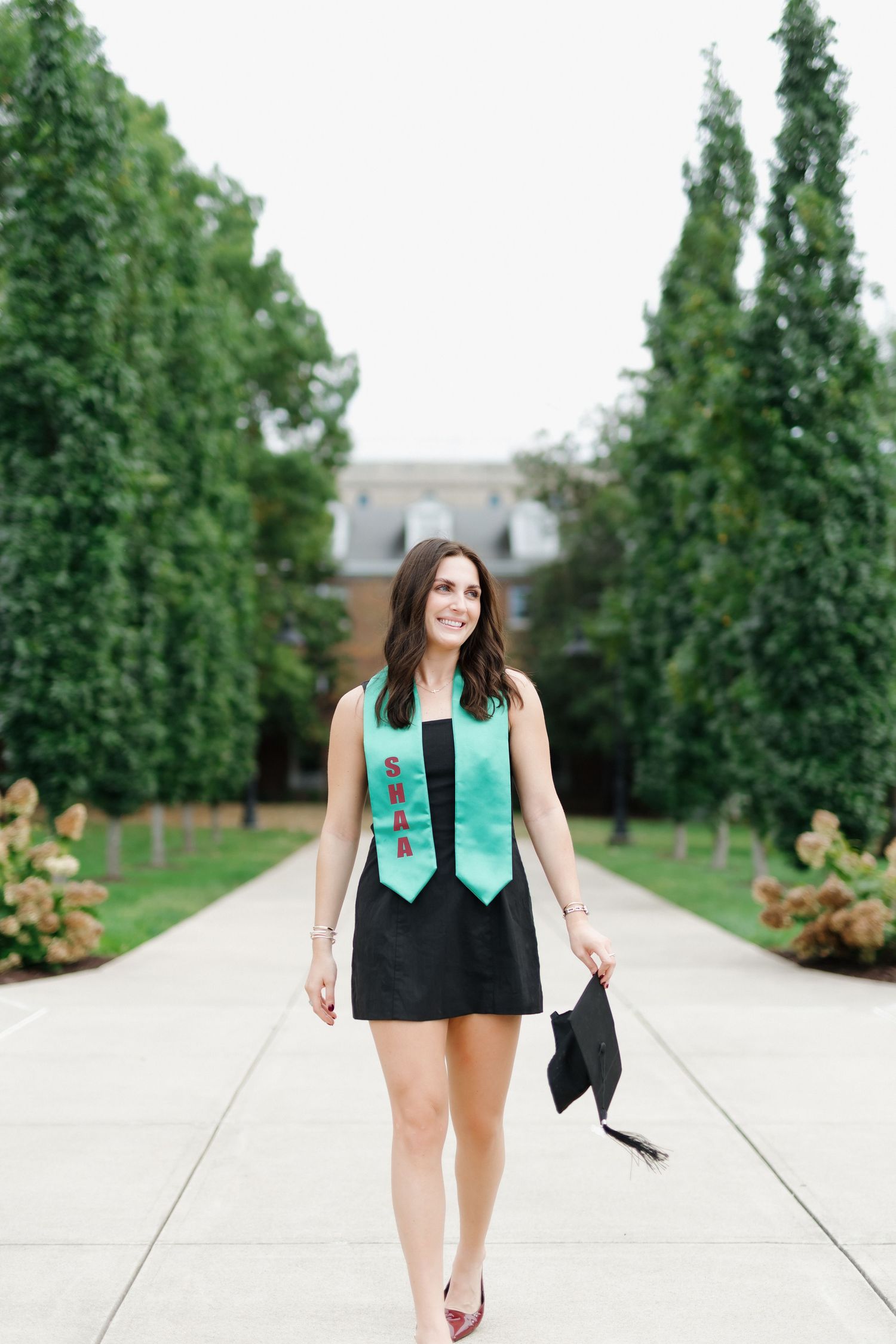 Smiling female graduate walks outdoors wearing black dress, teal graduation stole, holding cap on tree-lined campus path.