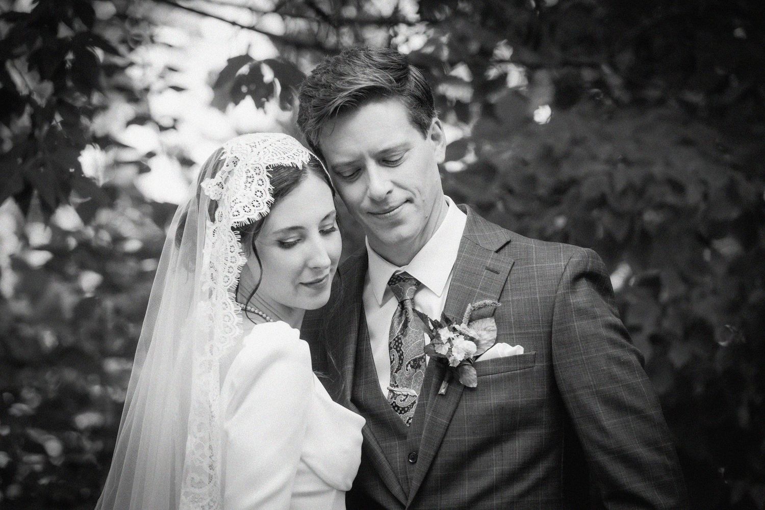 Black and white wedding photo of bride in lace veil and groom in plaid suit with boutonniere embracing outdoors.
