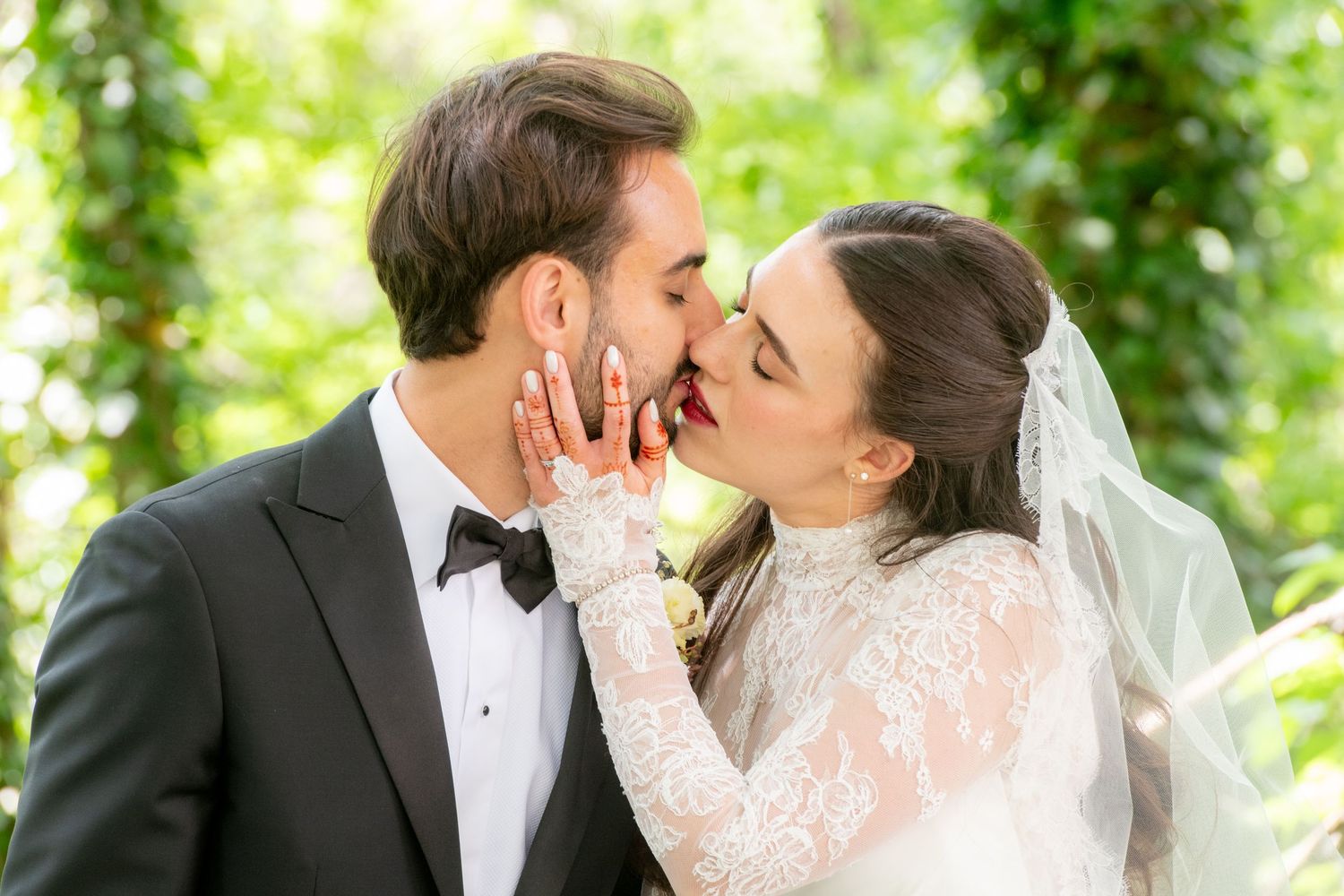 Bride and groom sharing a romantic kiss outdoors, bride wearing lace dress and veil, groom in black tuxedo.
