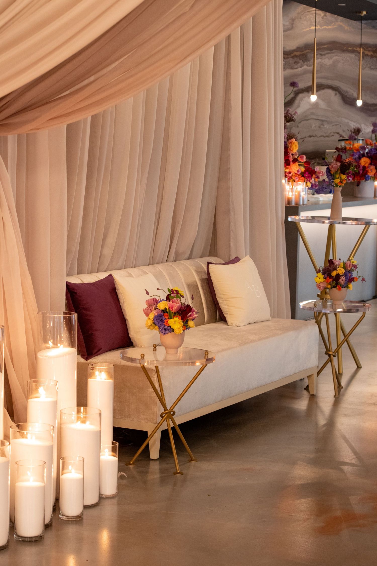Elegant wedding lounge with cream velvet sofa, burgundy pillows, gold tables, colorful floral arrangements, and glowing white pillar candles