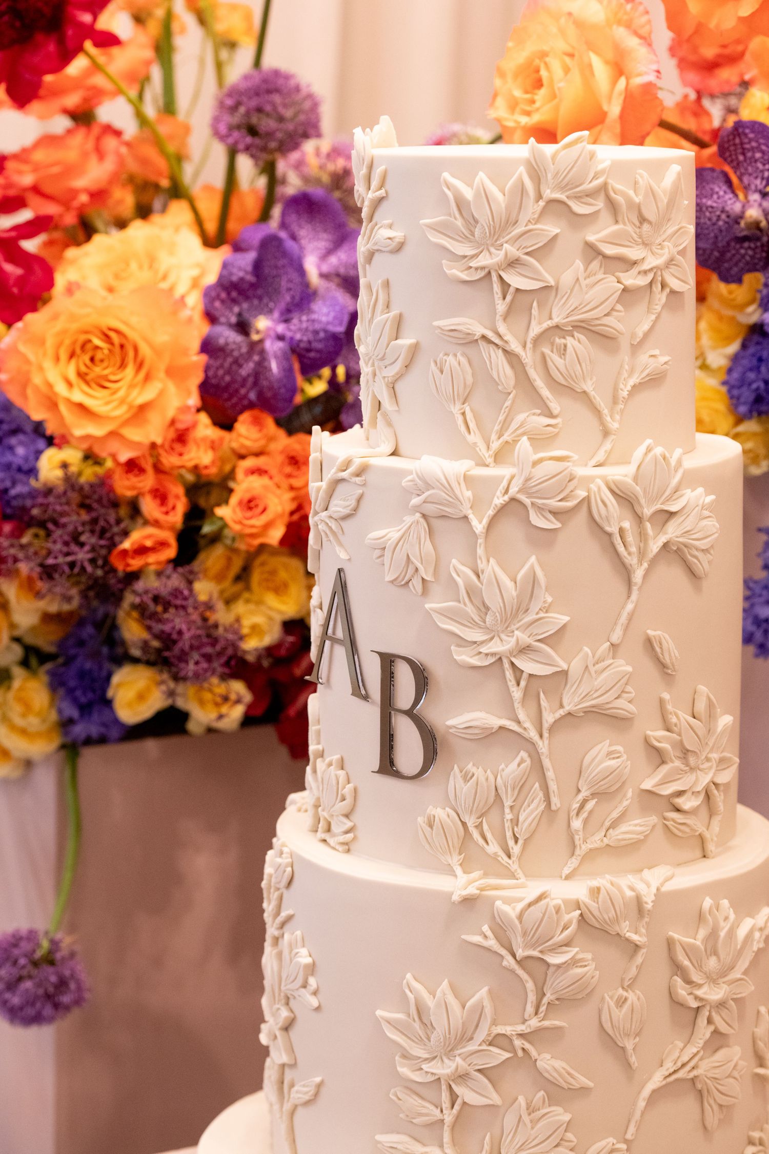 Elegant white three-tier wedding cake with floral relief design and AB monogram, set against vibrant colorful flower arrangements.