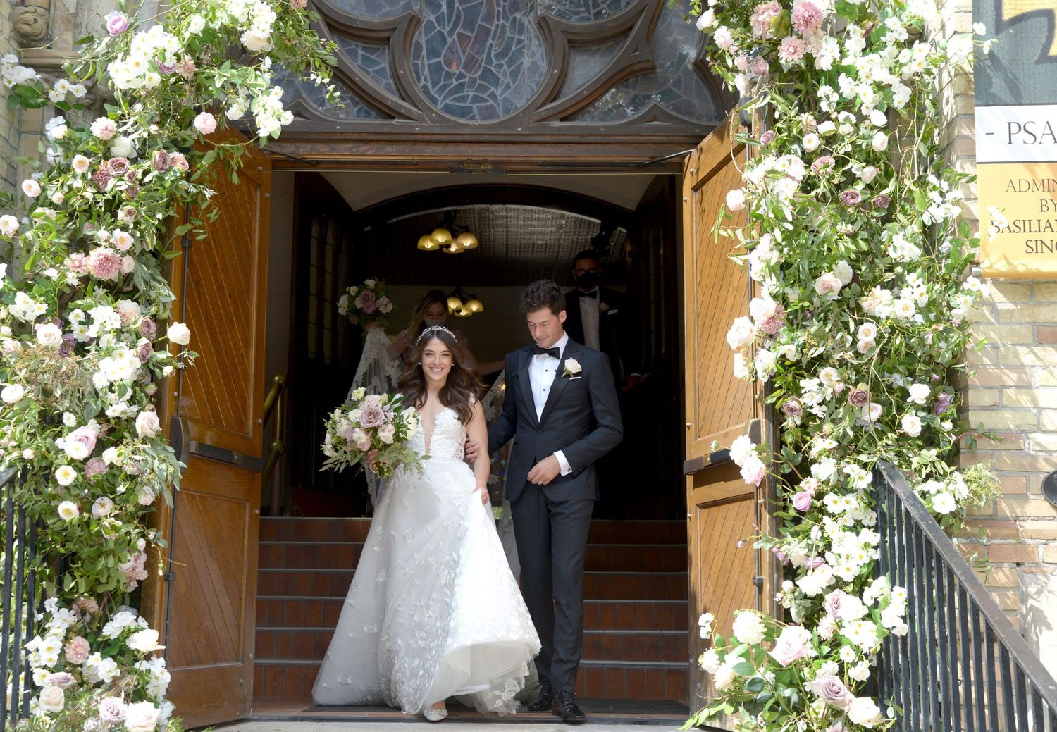 Newlyweds exit decorated church entrance under beautiful floral arch with white and pink blooms.