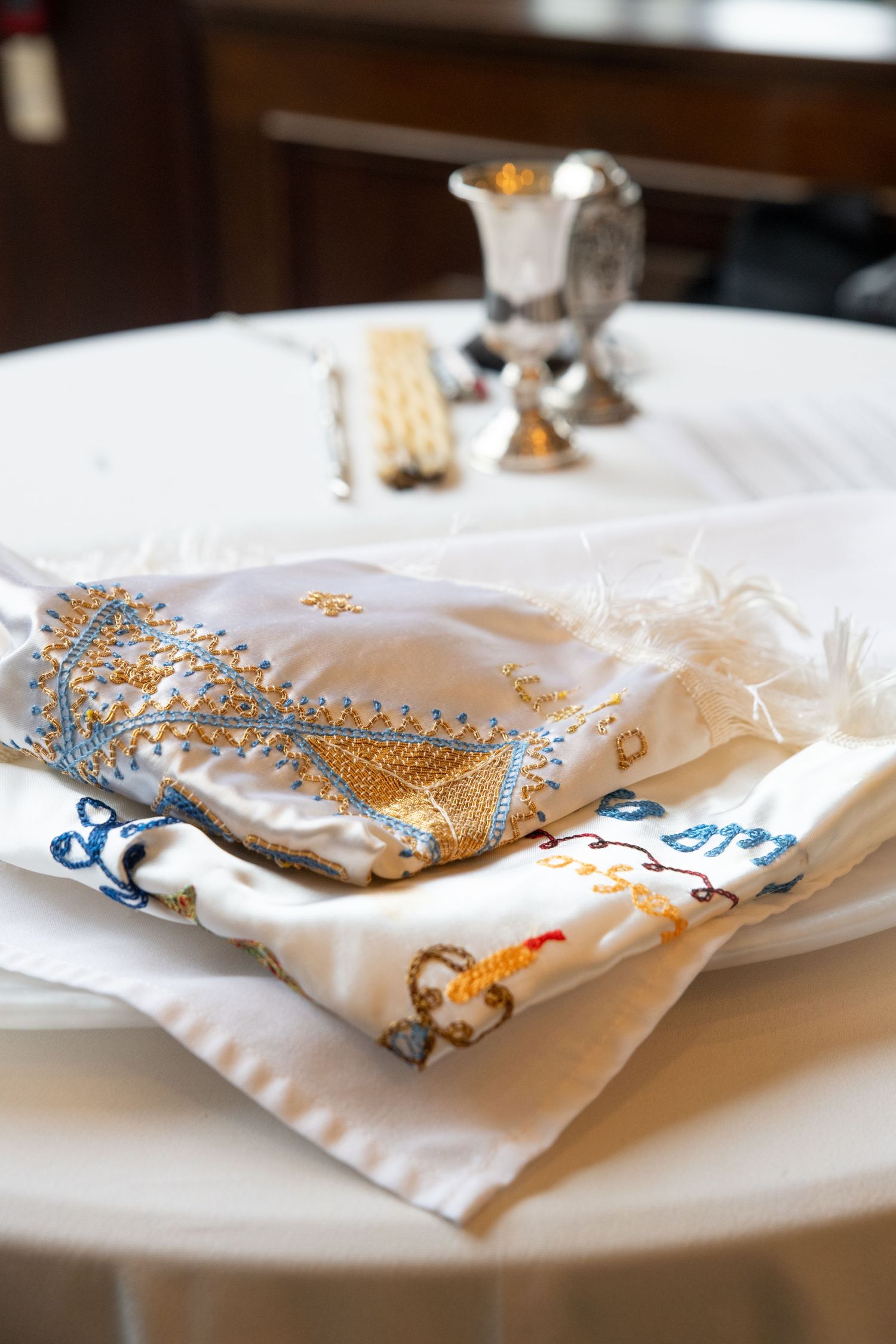Embroidered Jewish tallit prayer shawl folded on white table with silver Kiddush cup and matzah for Shabbat.