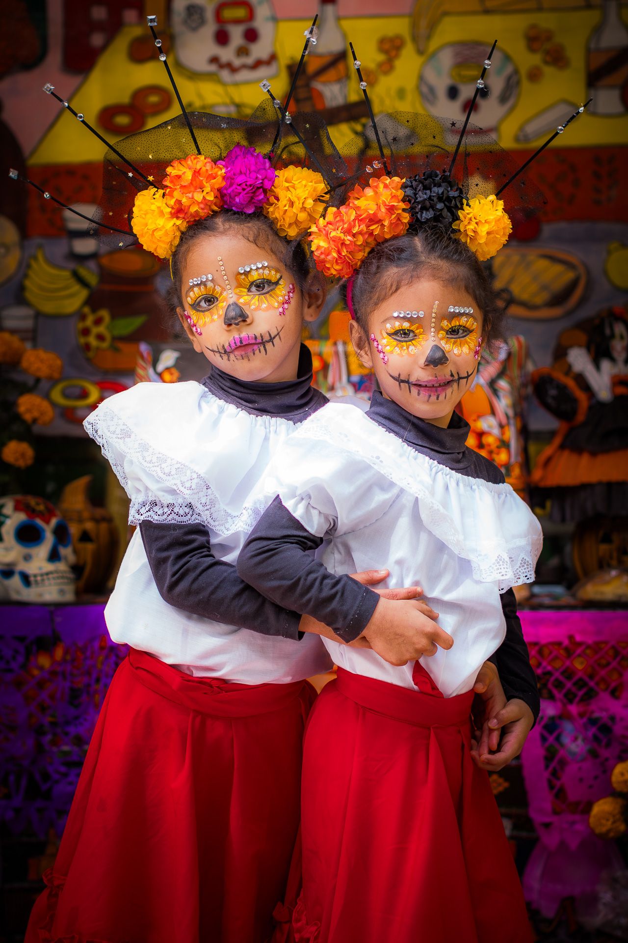 Two children in Day of the Dead face paint wearing white and red outfits with floral headpieces.