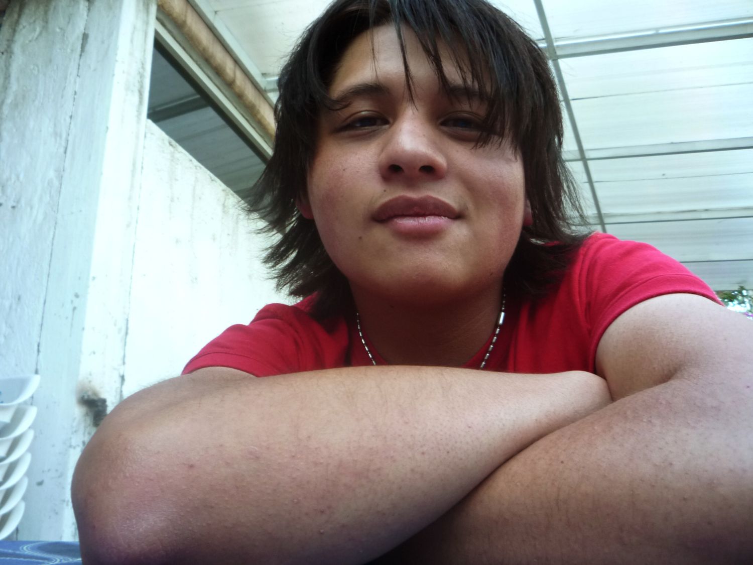 A teenager in a red t-shirt rests their arms on a surface and smiles at the camera in a selfie-style photo.