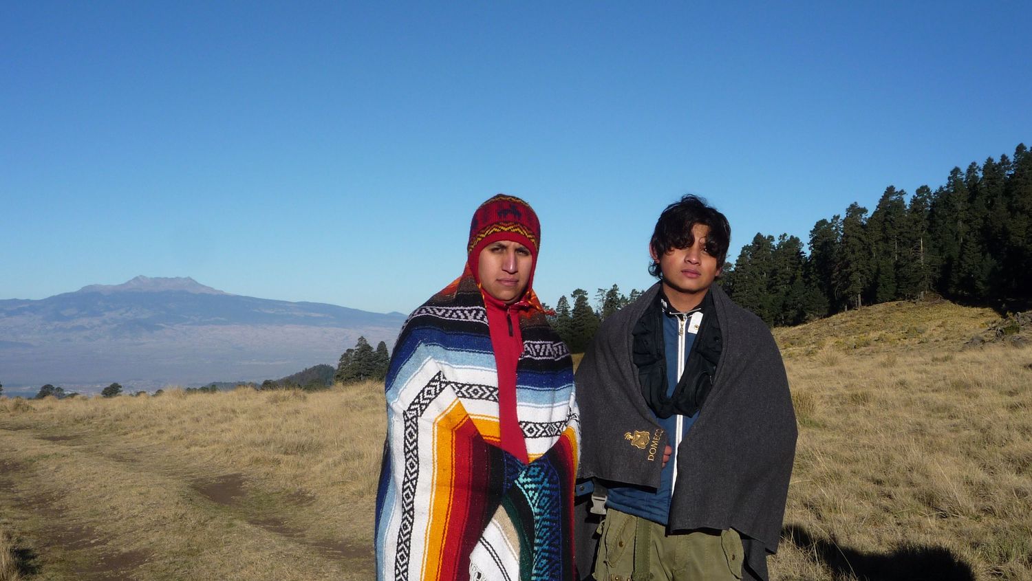 Two people stand together on a grassy hillside with mountains and forest visible in the background on a clear sunny day.