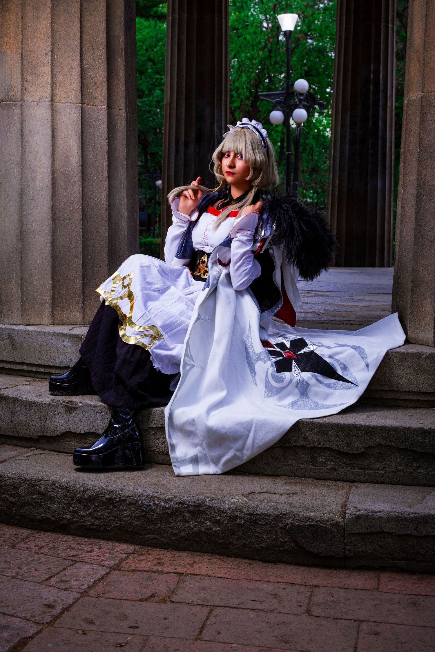 Cosplayer in an ornate white and black dress costume poses elegantly on stone steps near classical columns and lamp posts.