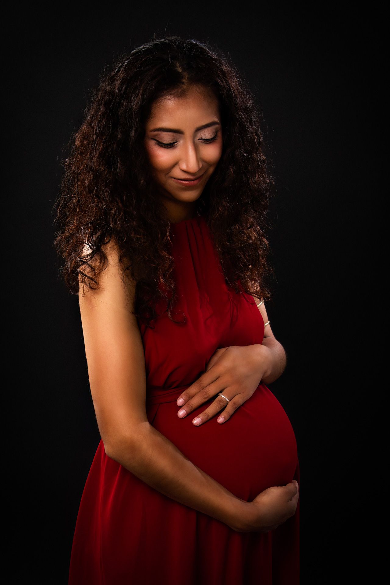 Expectant mother in red dress poses against dark background in maternity photoshoot.