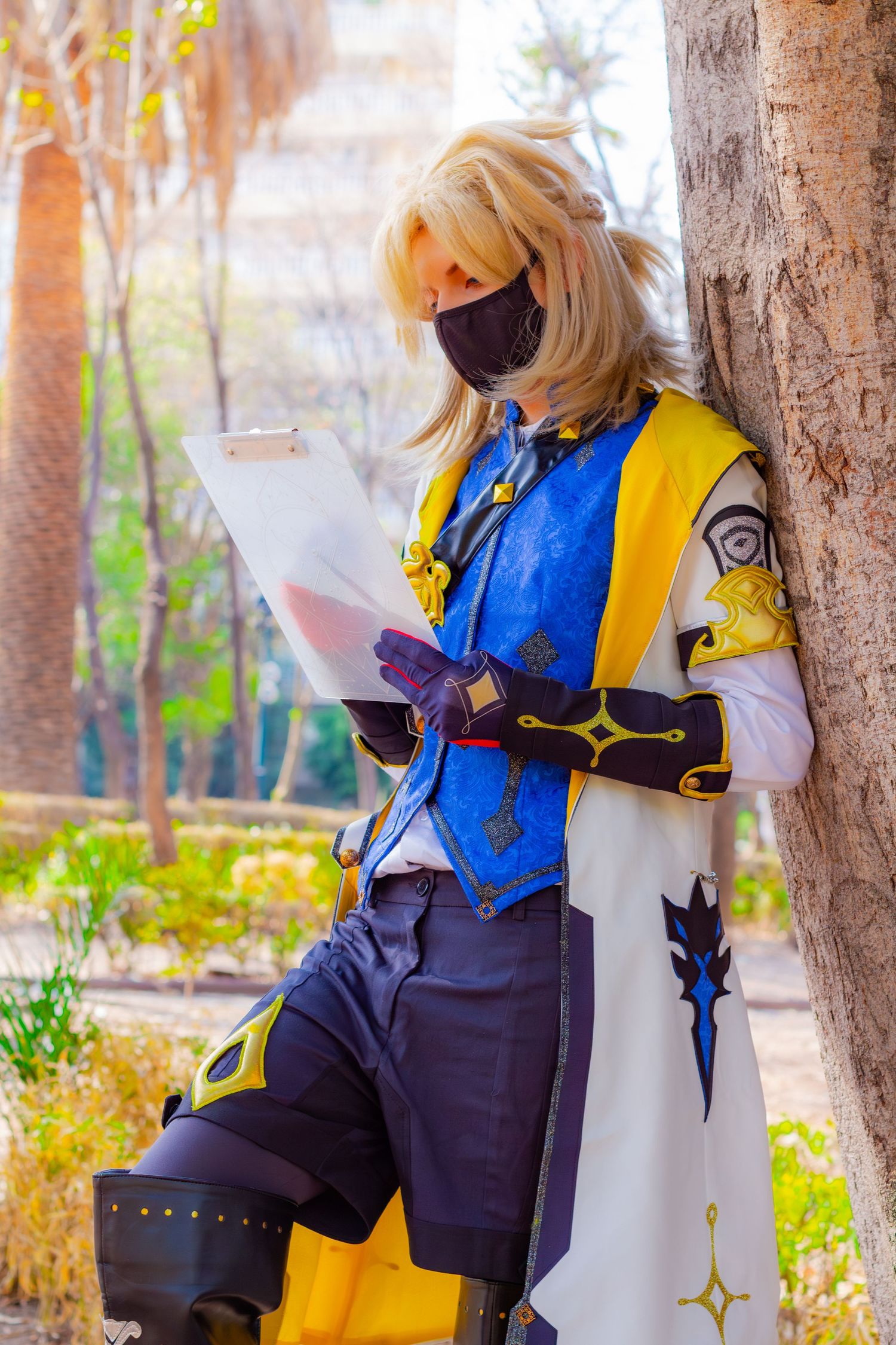 Cosplayer in blue and yellow fantasy outfit reading scroll in outdoor garden setting.