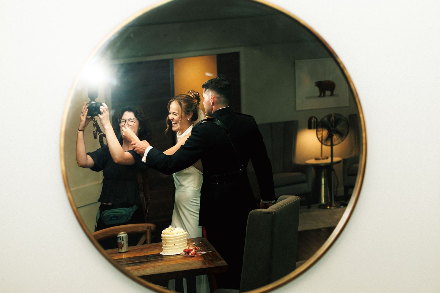 Couple sharing a joyful moment reflected in a round mirror, captured by a photographer at their wedding reception.