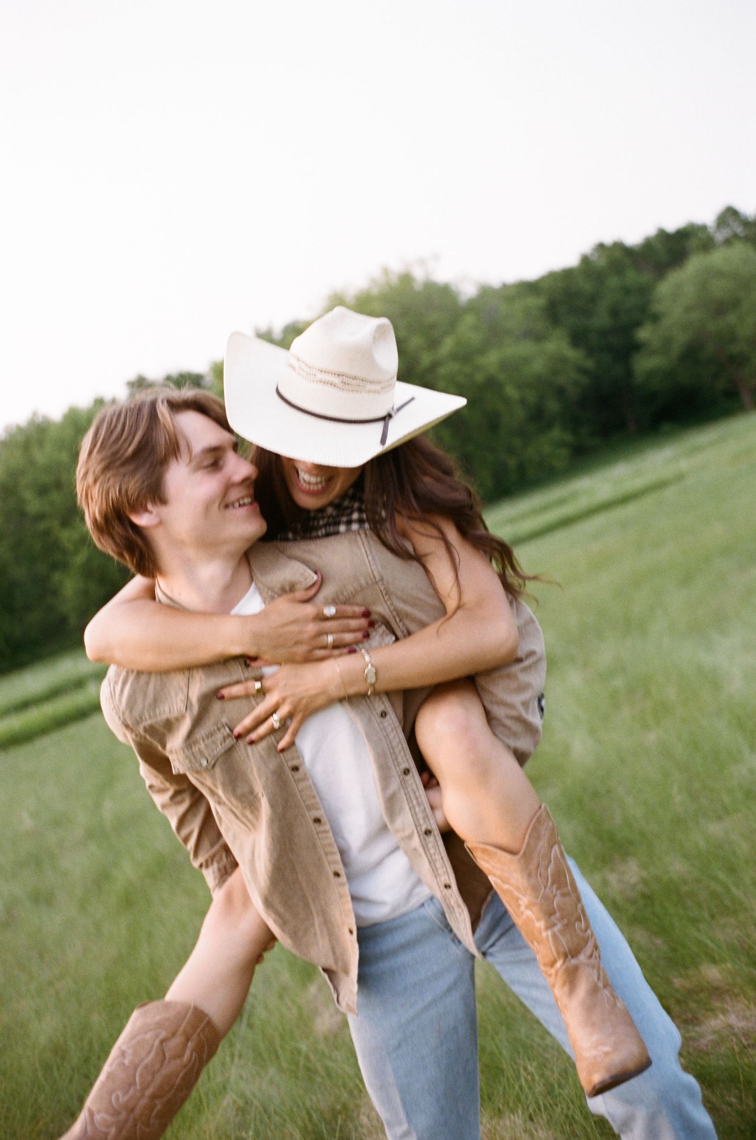 A couple embraces romantically in a green meadow on a sunny day, with one wearing a cowboy hat and denim.
