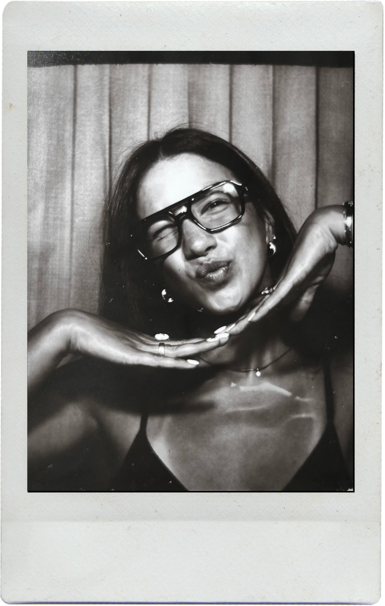 A black and white Polaroid photo shows someone playfully posing in glasses against a curtained background.