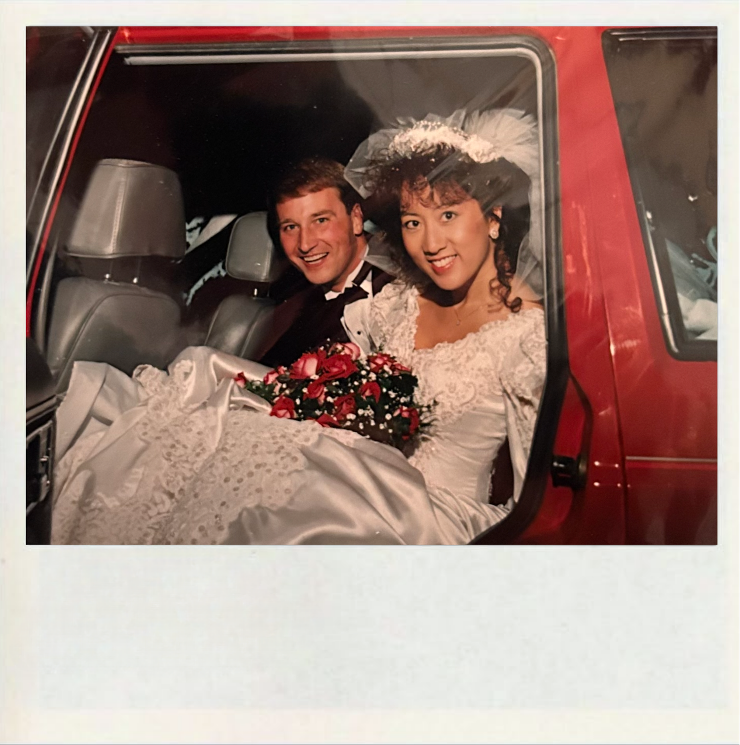 A couple in wedding attire sitting in a vintage red vehicle decorated with flowers and ribbons.