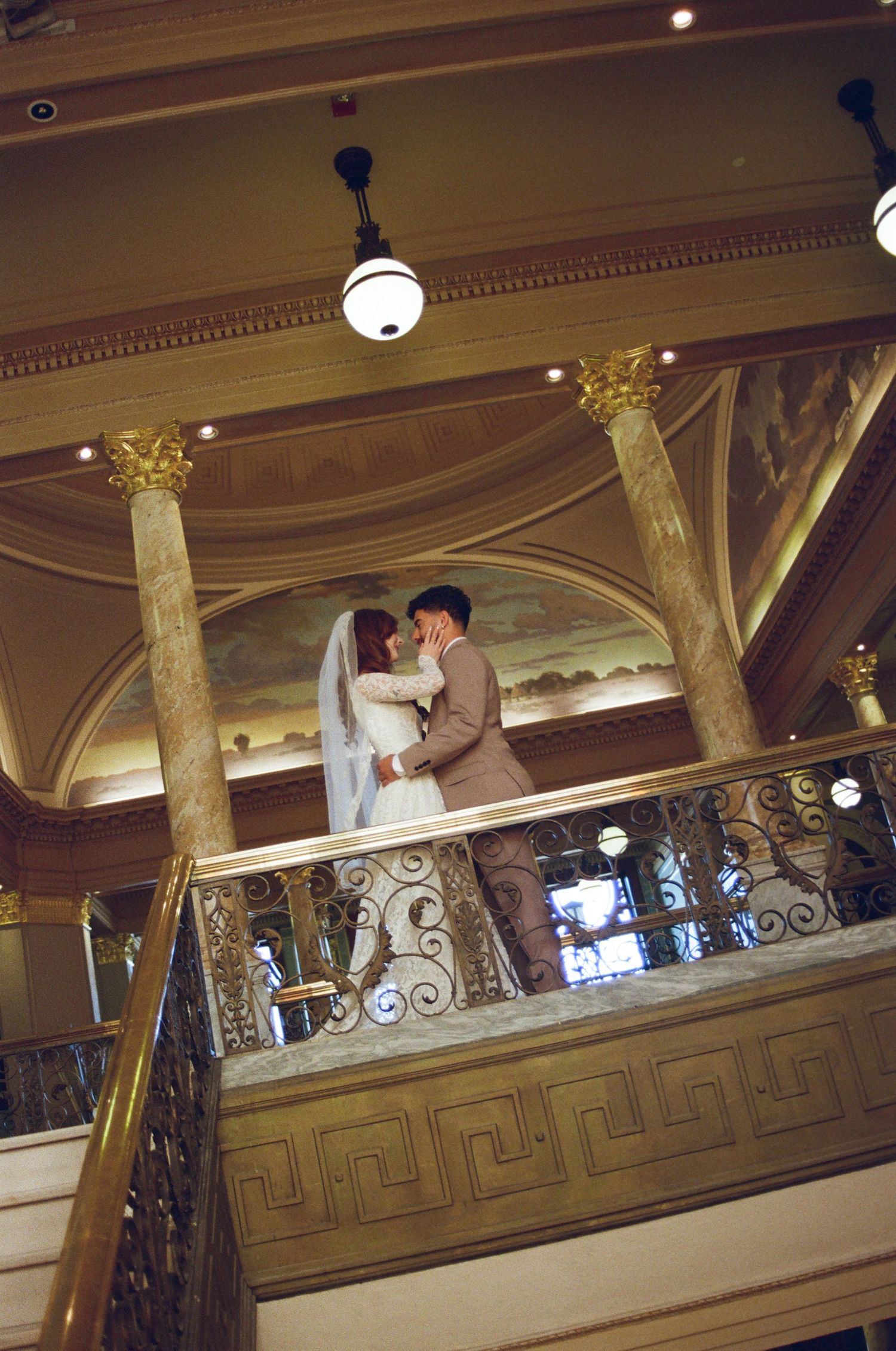 A couple shares a romantic moment on an ornate balcony with gold columns and decorative railings in a grand ballroom.