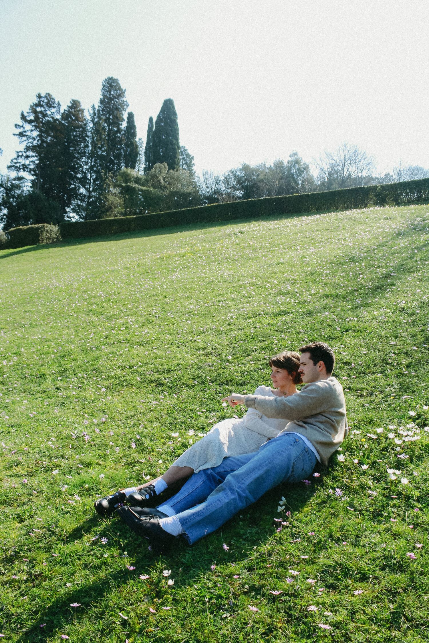 Two people relax together on a grassy slope with trees in the background.