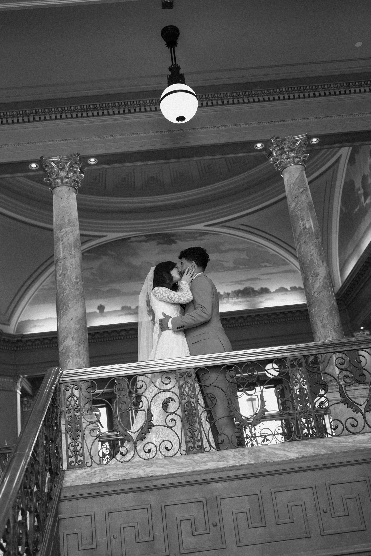 A couple shares a romantic moment on an ornate balcony with decorative railings in a black and white photograph.