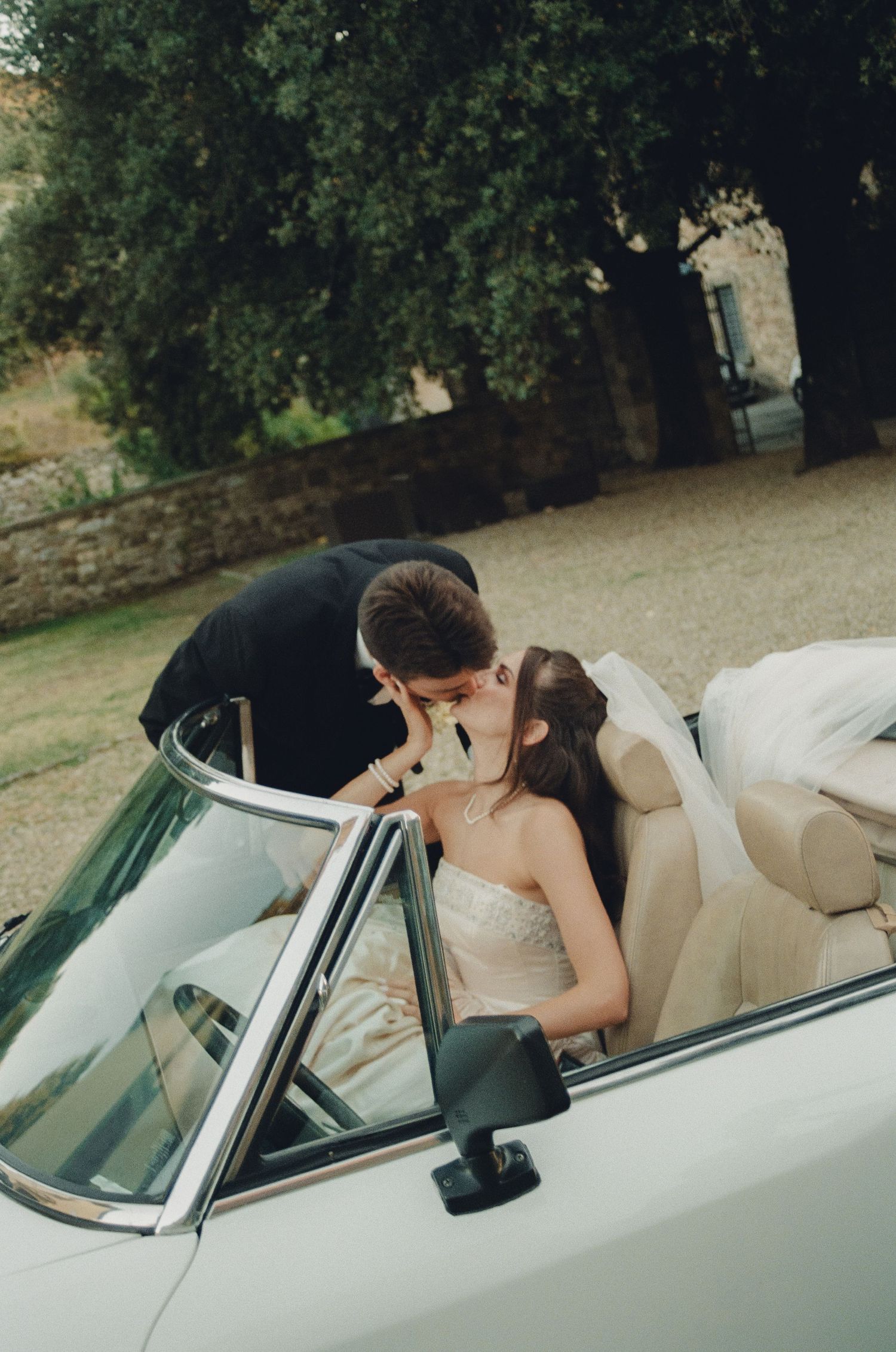 A romantic moment captured in a vintage white convertible car parked under trees.