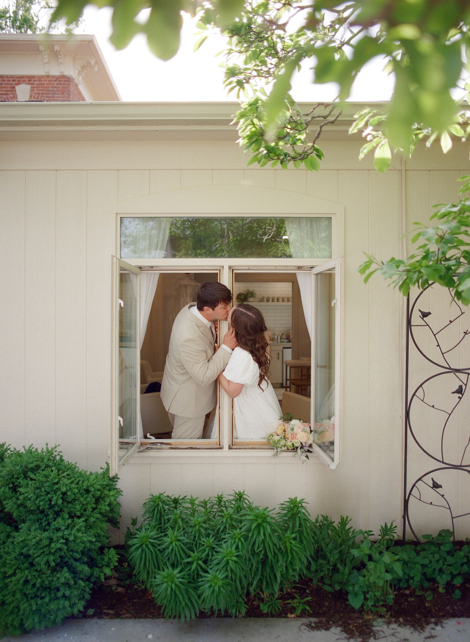 Bride and groom share a romantic kiss framed in an open window surrounded by lush greenery on their wedding day.