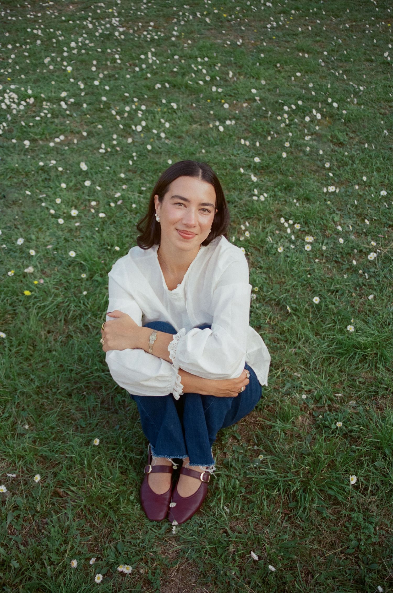 Someone in a white sweater and blue jeans sits cross-legged on grass scattered with small white flower petals.