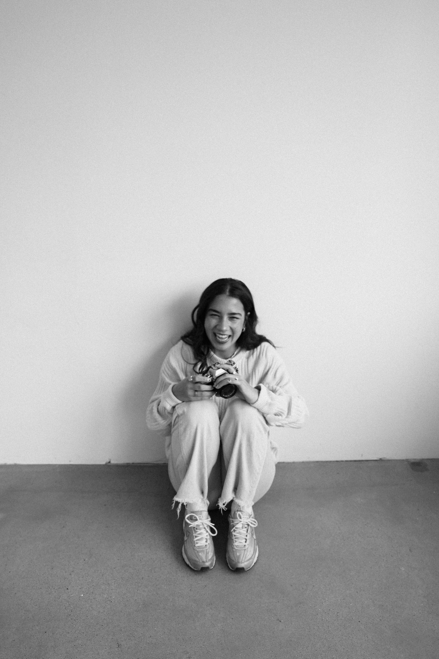 Person in white sweatshirt and sneakers sits casually against white wall.