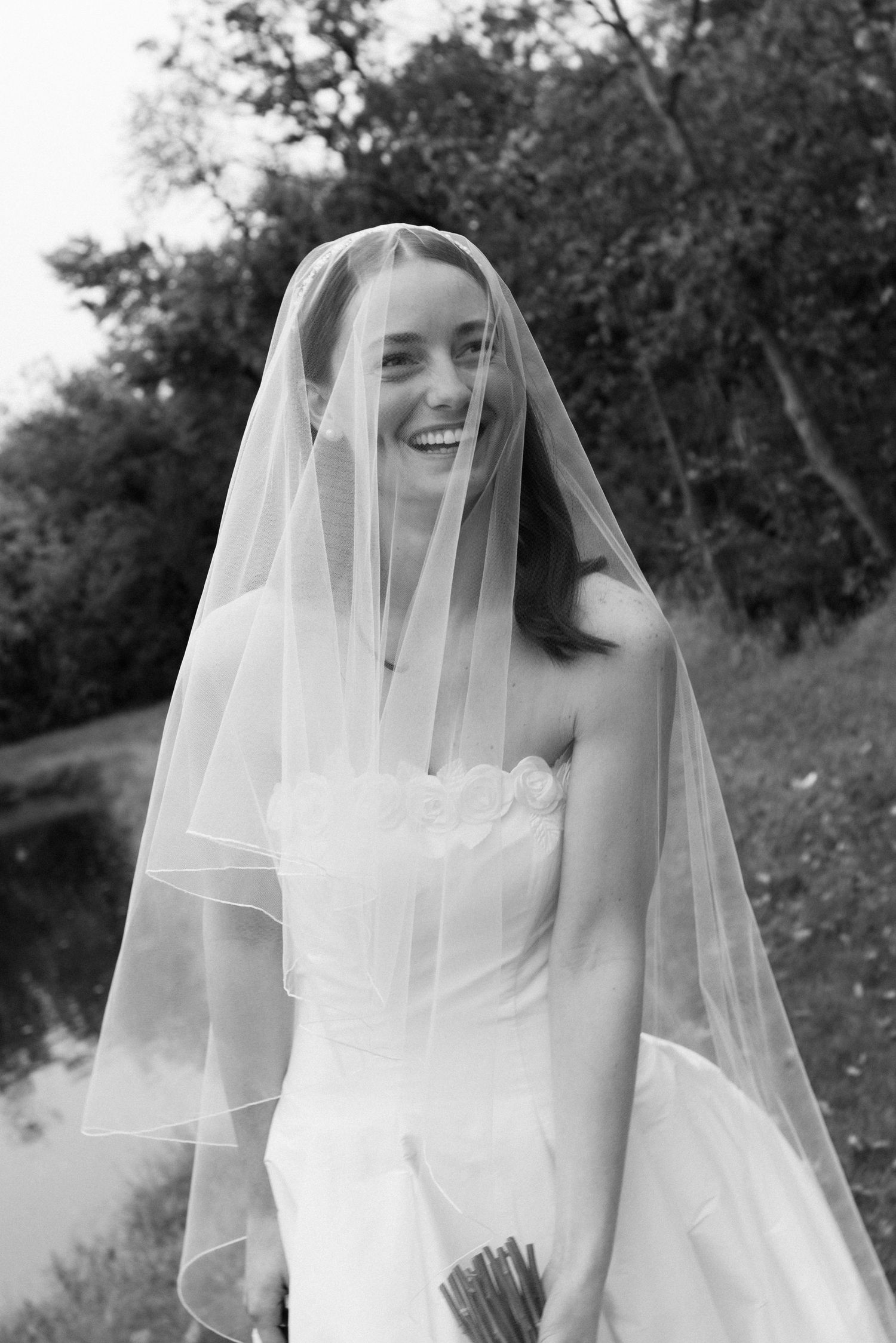 A joyful bride in a classic white wedding dress and veil poses outdoors in a black and white photograph.