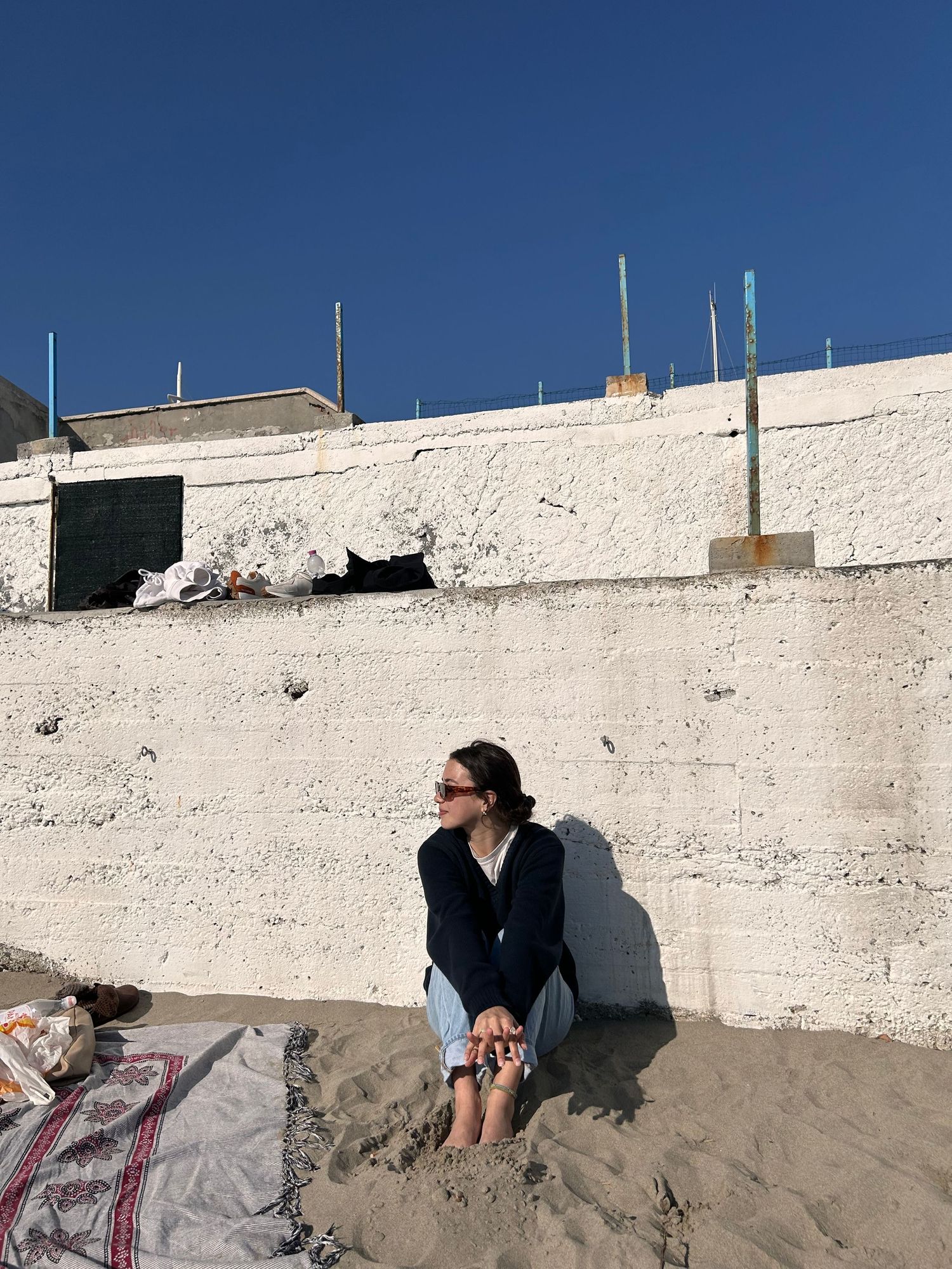 Person sitting against white wall at beach on blanket wearing sunglasses and casual clothing.