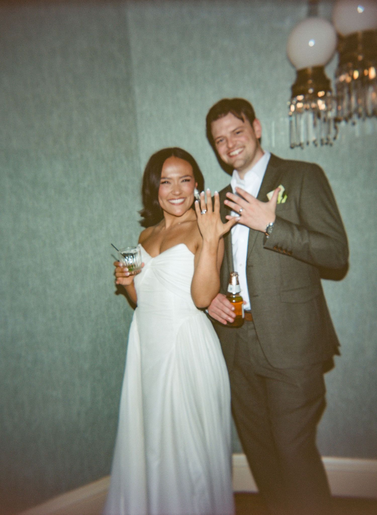 Smiling bride in white gown and groom in gray suit showing wedding rings while holding drinks at their reception.