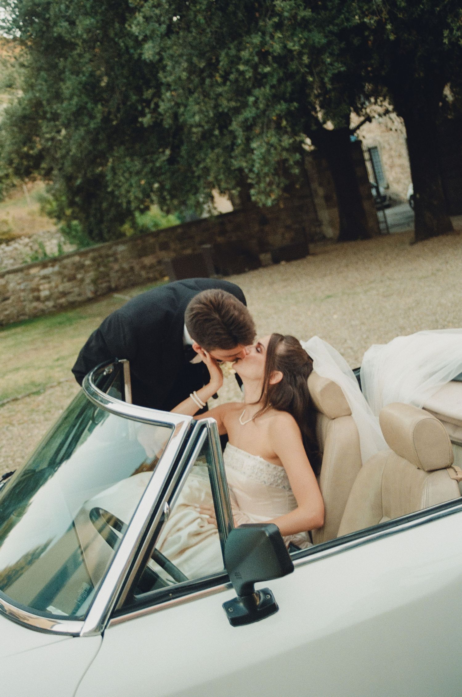 A couple shares romantic moments in a vintage white convertible car parked under trees.