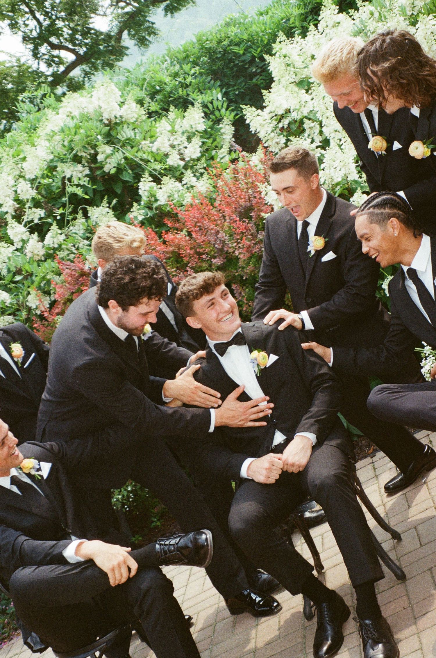 A sequence of groomsmen in black suits playfully pushing and falling into each other in a domino effect outdoors.