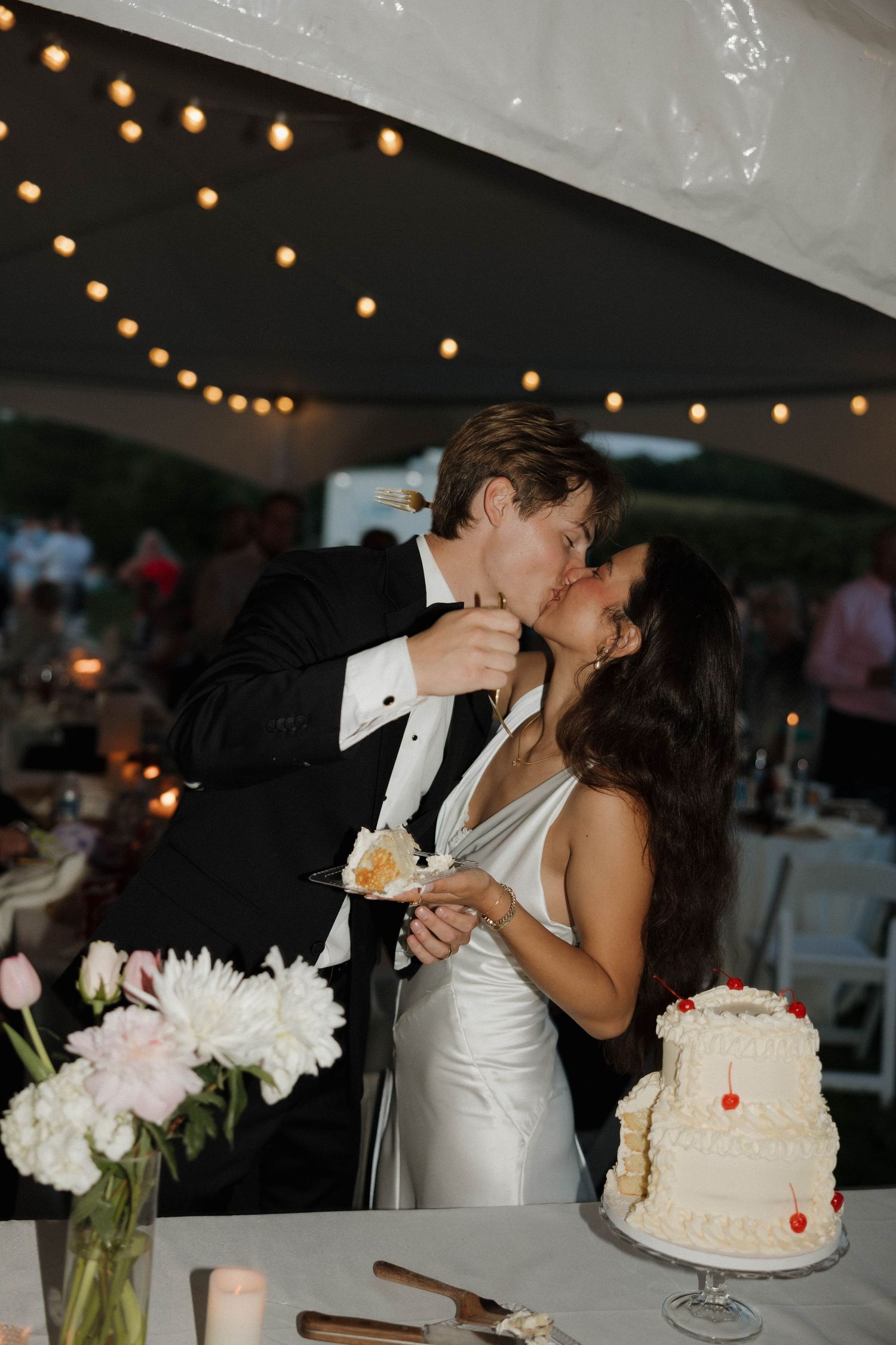 A romantic wedding moment with string lights overhead as newlyweds share cake at their reception.