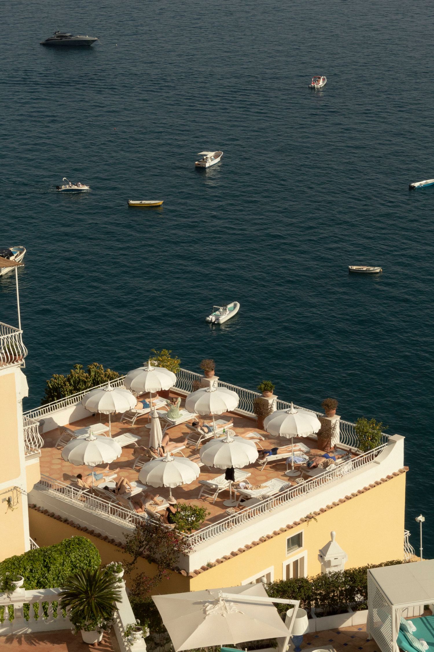 Rooftop terrace with white umbrellas overlooking boats on deep blue Mediterranean waters.