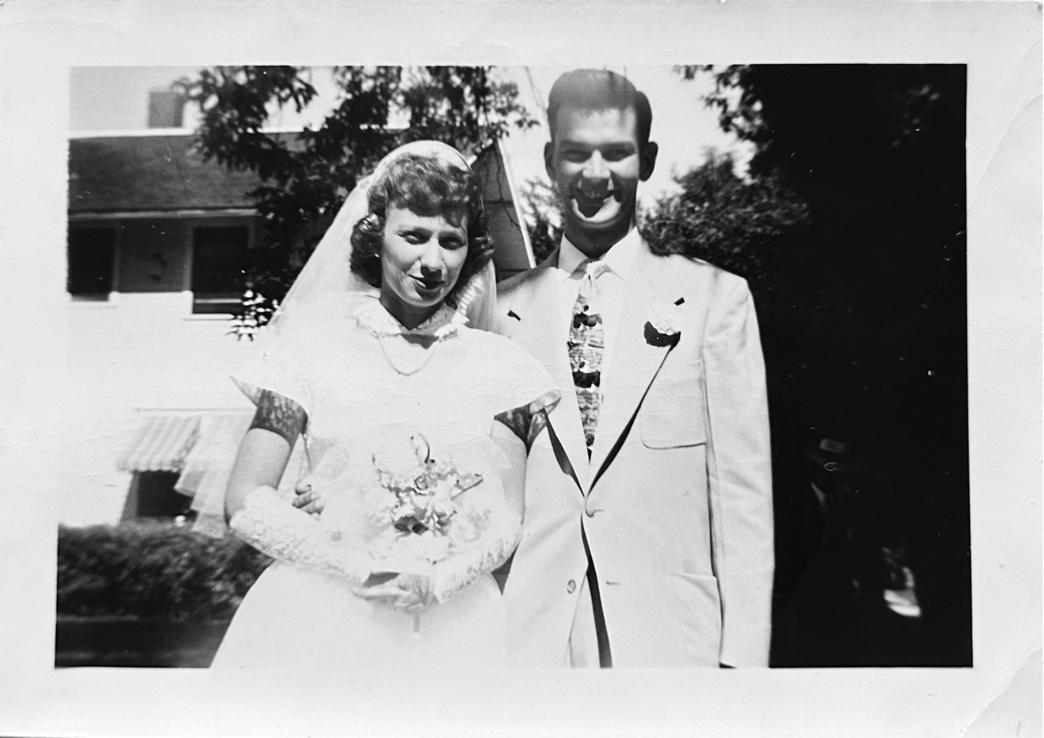 A vintage black and white photograph shows a couple in formal attire, with the woman holding a bouquet of flowers.