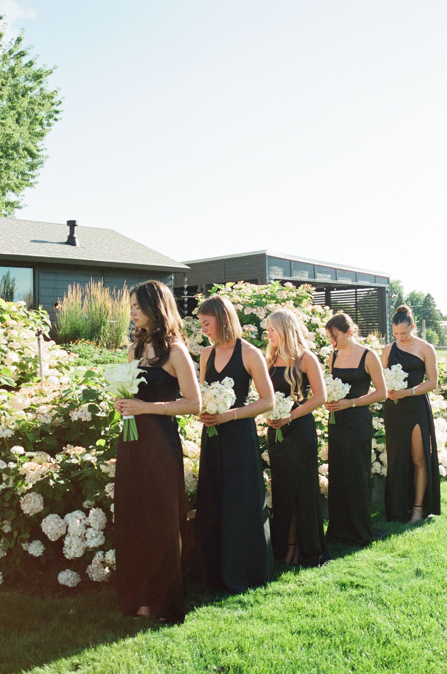 A row of bridesmaids in black dresses stand along a floral-decorated wall at an outdoor wedding venue.