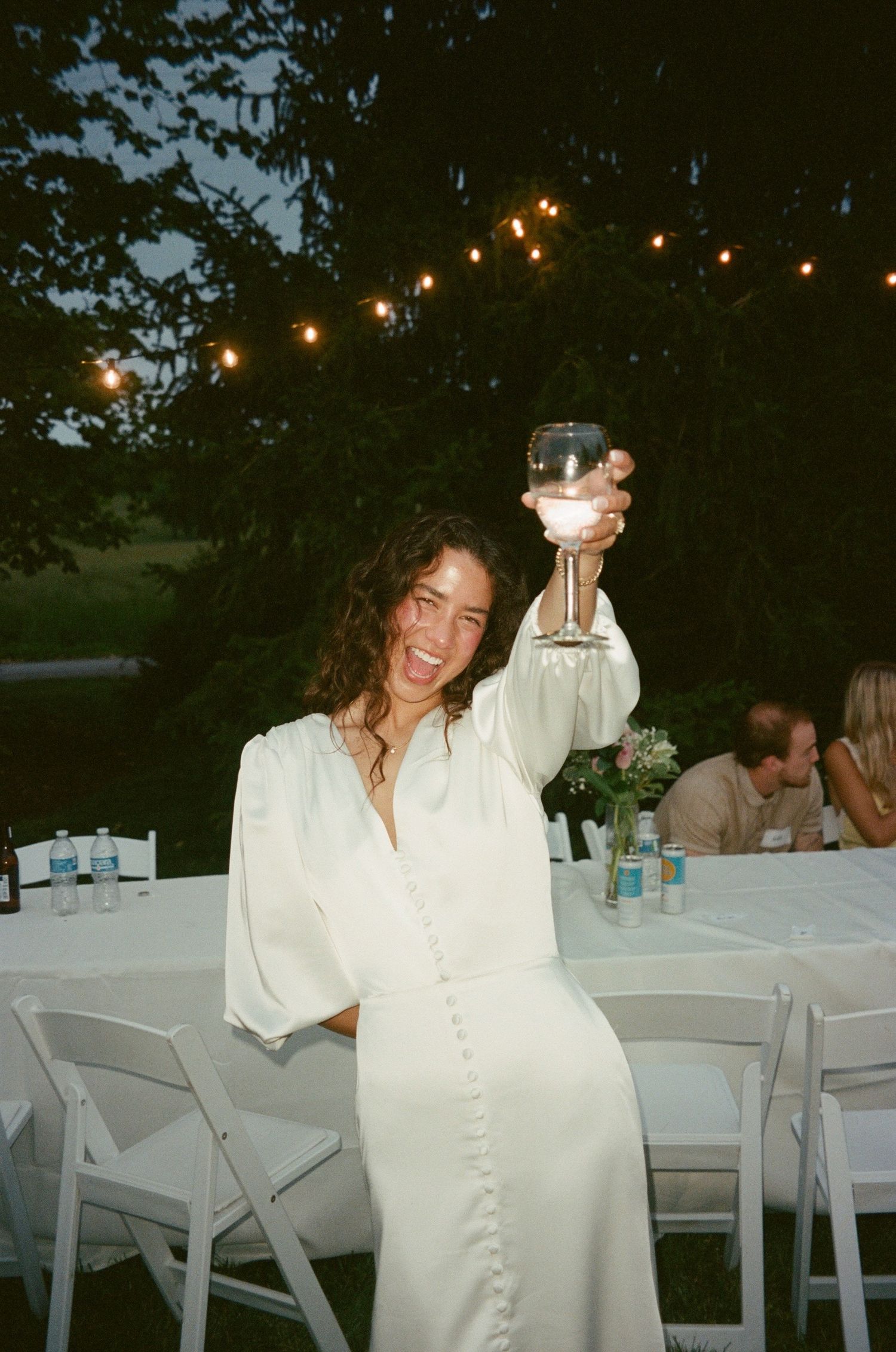 Wedding guest in white dress raising glass during outdoor reception with string lights.