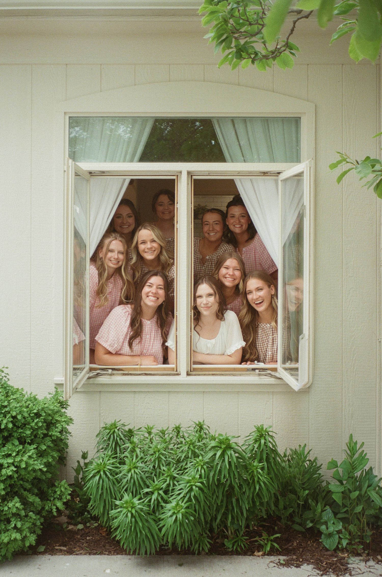 Group of smiling women peeking through an open white window surrounded by lush green shrubs and curtains.
