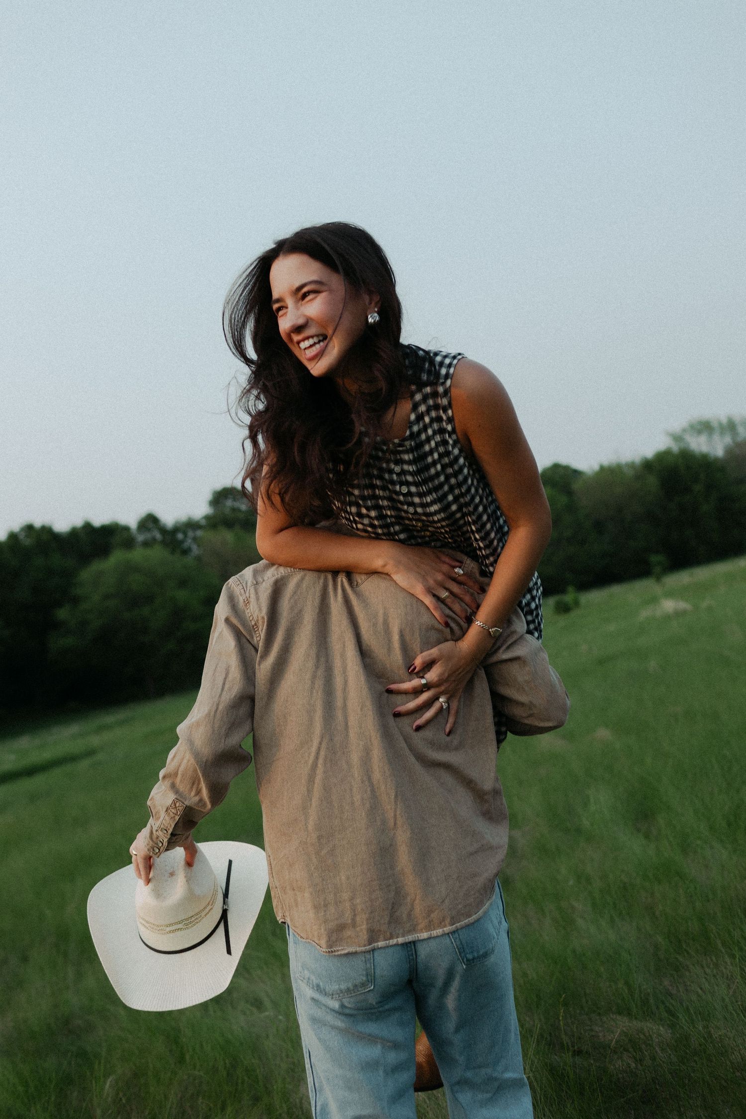 A couple embraces and laughs together in a grassy field on a cloudy day.