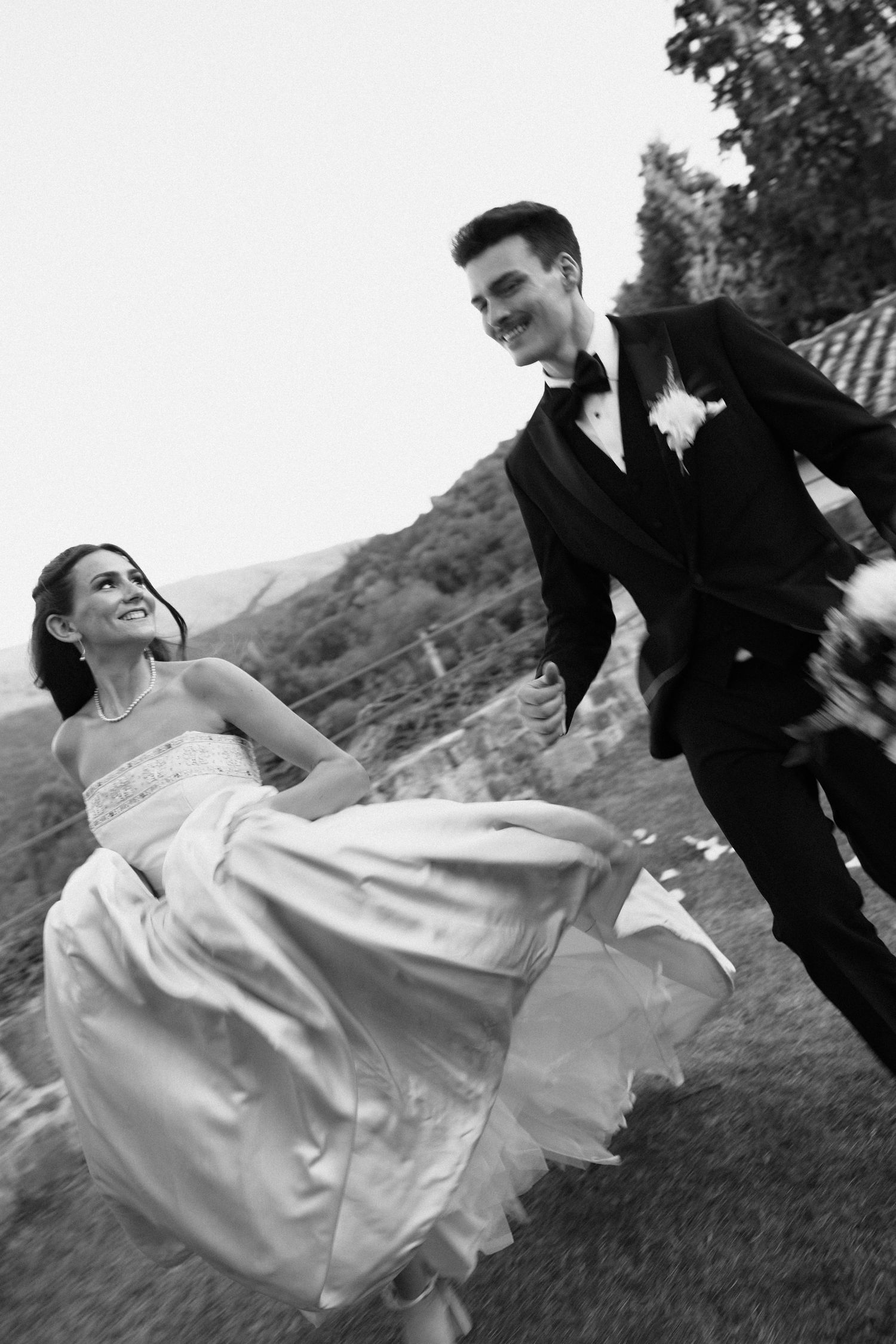 A couple in wedding attire playfully dances and spins on a grassy hillside in black and white.