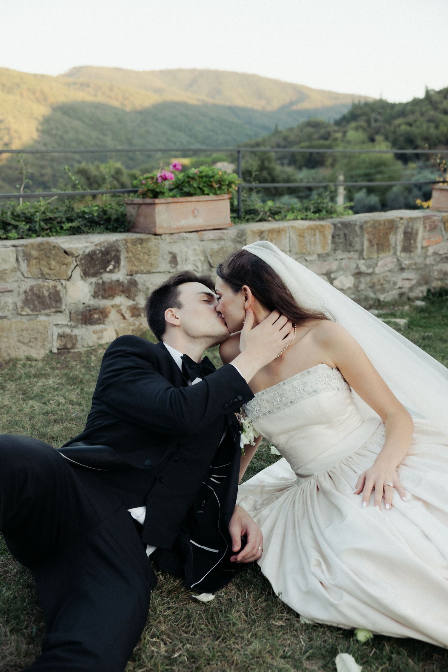 A romantic wedding couple shares intimate kisses while sitting on a stone wall with mountains in the background.