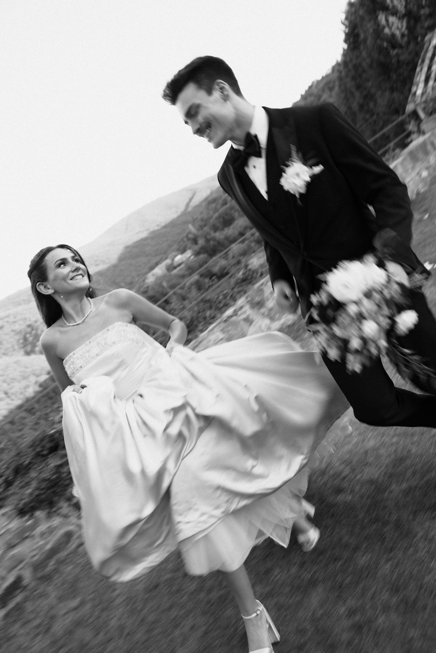 A couple in wedding attire dances playfully in a field with mountains in the background, captured in black and white.