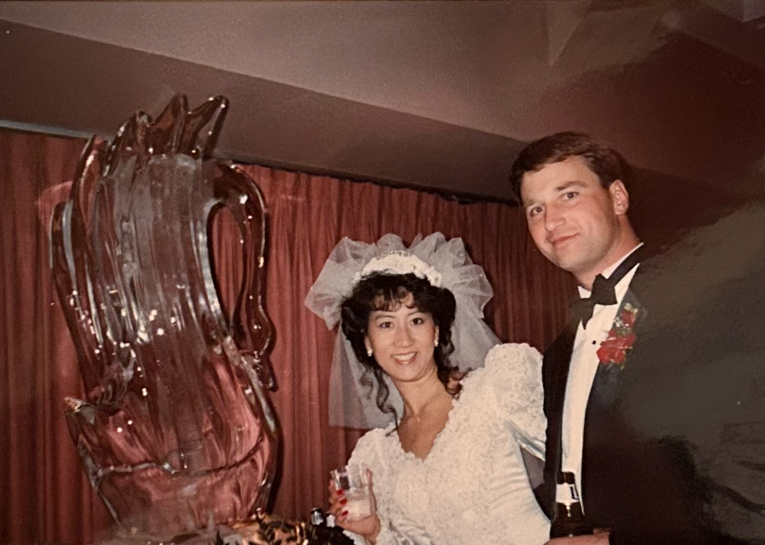 A wedding couple poses next to a large ice sculpture at their reception in vintage-style photograph.