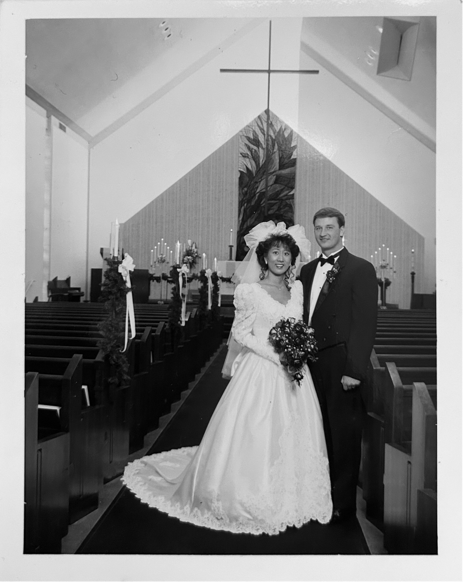 A couple in formal wedding attire poses together in a church sanctuary with wooden pews and a cross visible behind them.