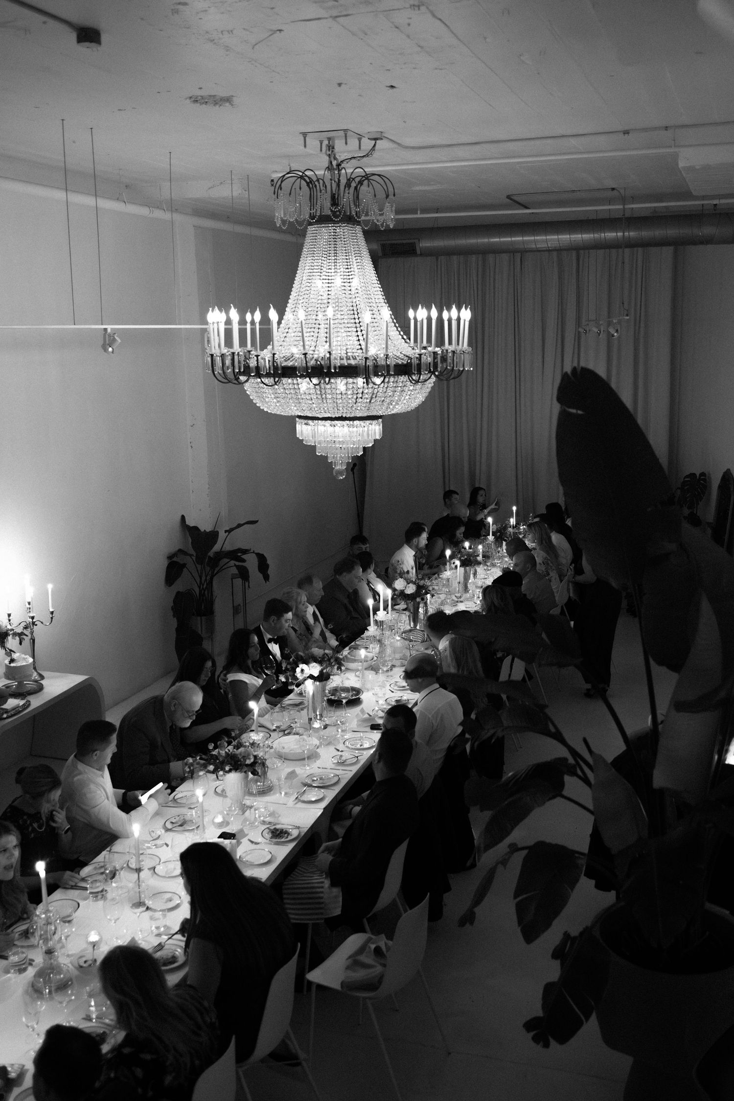 A formal dinner party under an elegant crystal chandelier with guests seated at a long table in black and white.