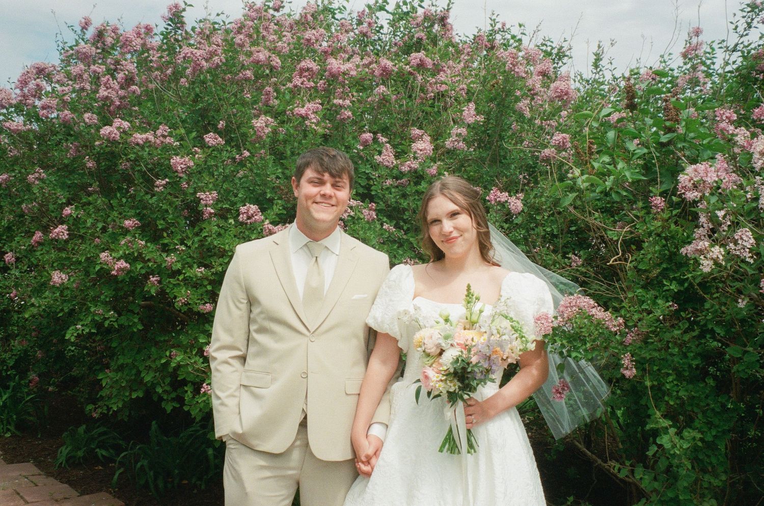 Bride and groom posing together in front of blooming pink flowering bushes, bride holding a pastel bouquet.