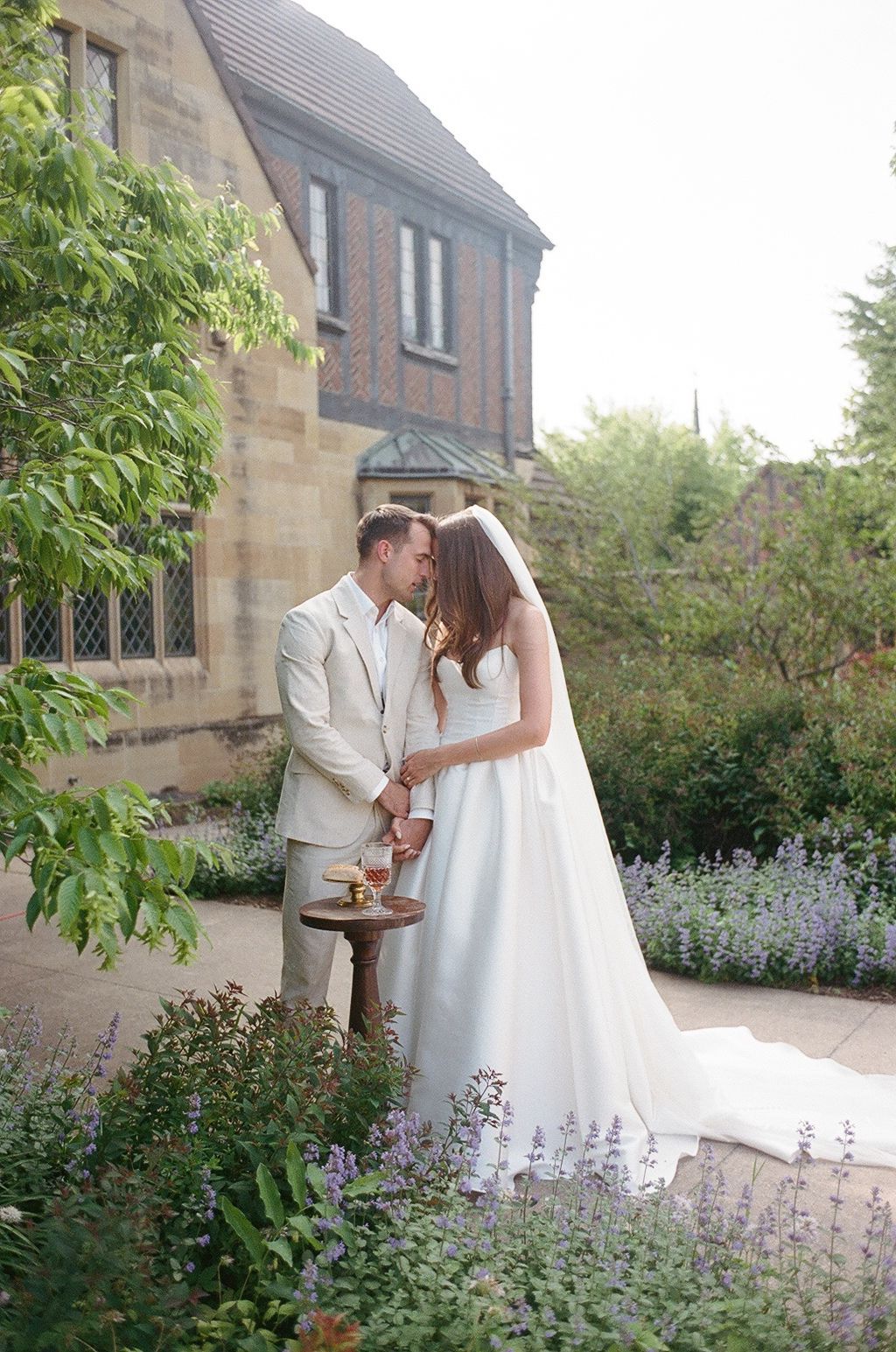 A romantic moment between a couple in white attire standing among blooming lavender flowers outside a stone building.