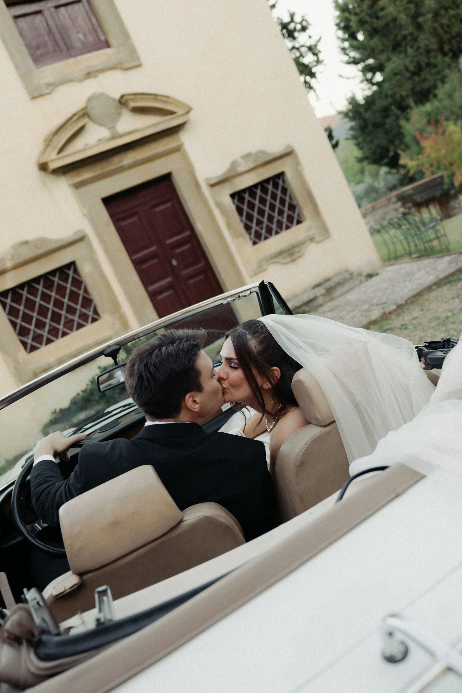 A couple shares a romantic moment in the back seat of a vintage cream-colored convertible car outside a historic building.